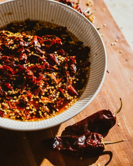 Overhead shot of a white bowl with red chili flakes and seeds, along with two dried chili peppers on a wood surface.