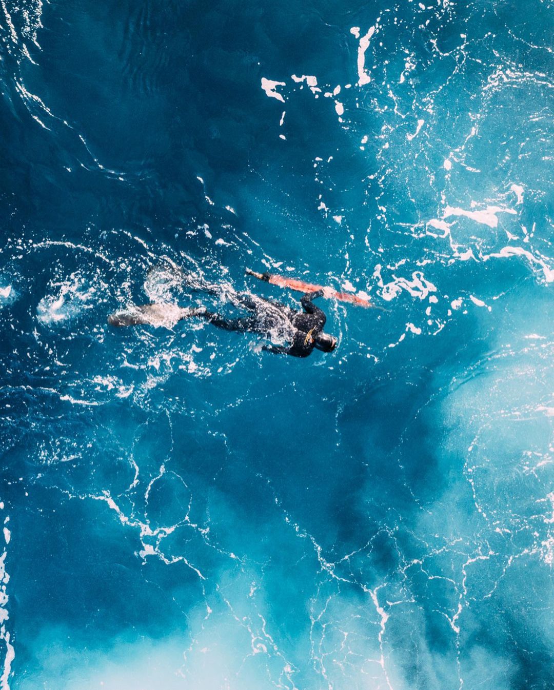 Aerial view of a person swimming in a dark wetsuit alongside a surfboard in vividly patterned deep blue and turquoise ocean water.
