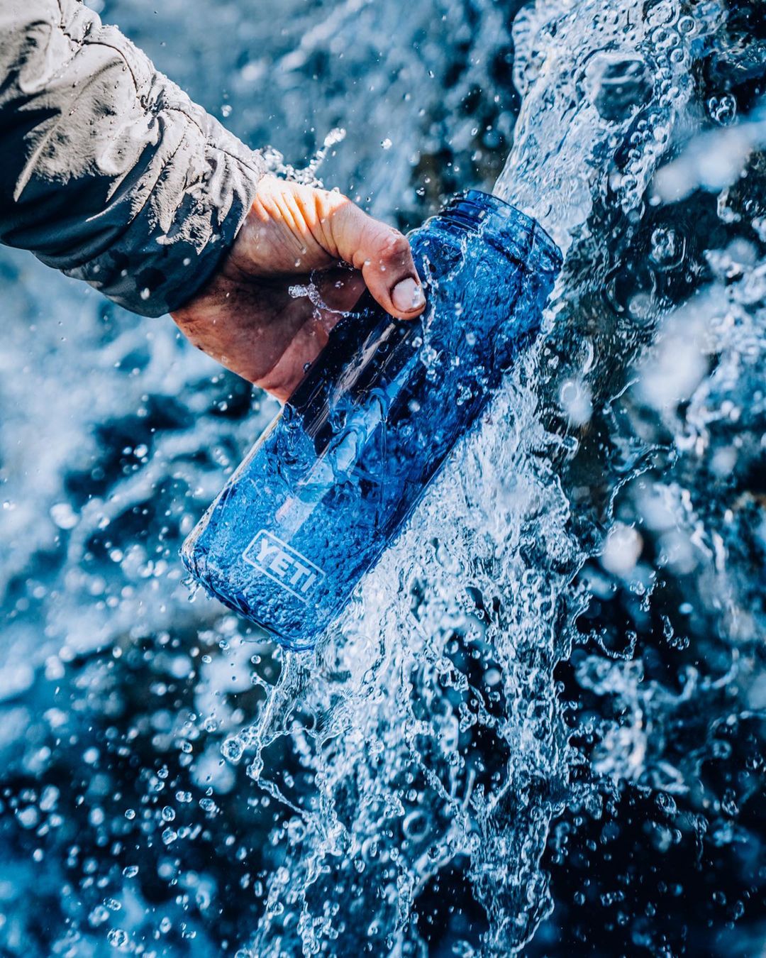 Hand holding a blue Yeti water bottle with water cascading around it, close-up shot of refreshment in an outdoor setting.