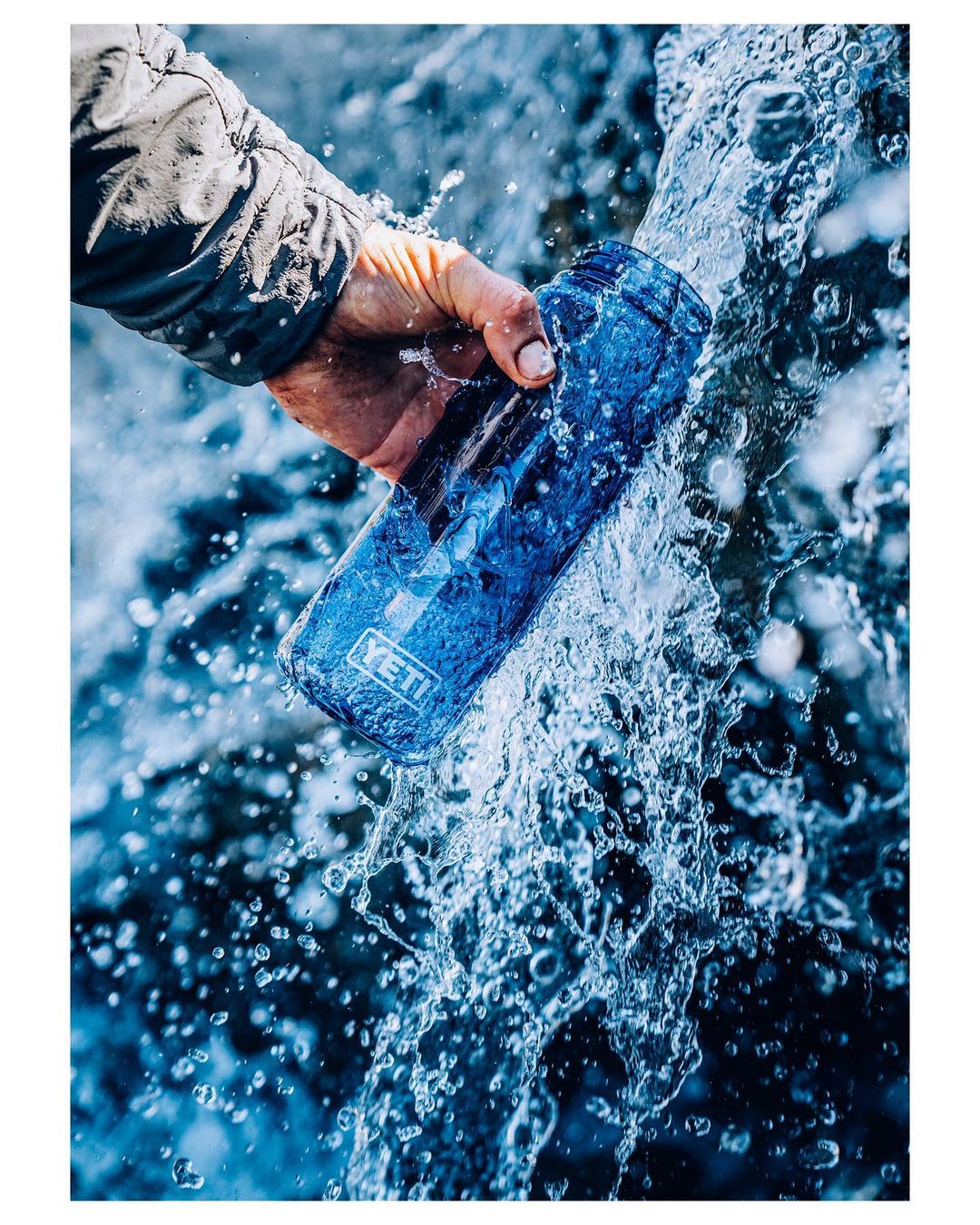 An arm holds a blue YETI water bottle under a waterfall with clear water splashing against the bottle and the person's hand.