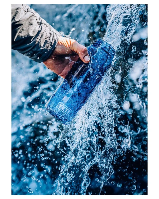 An arm holds a blue YETI water bottle under a waterfall with clear water splashing against the bottle and the person's hand.