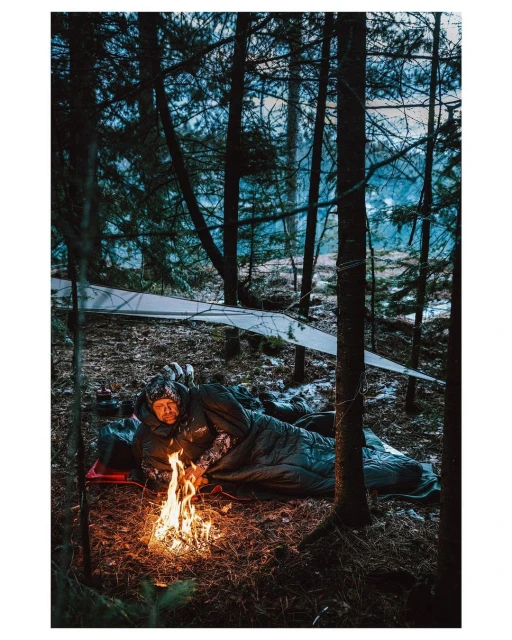 Adult male in sleeping bag near campfire in forest environment under a tarp shelter, framed by tall trees in an outdoor environment.