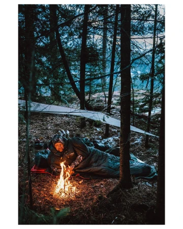 Adult male in sleeping bag near campfire in forest environment under a tarp shelter, framed by tall trees in an outdoor environment.