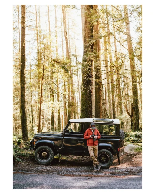 Adult male in orange jacket stands near a black Land Rover in a dense forest with tall trees on a sunny day in a scenic outdoor shot.