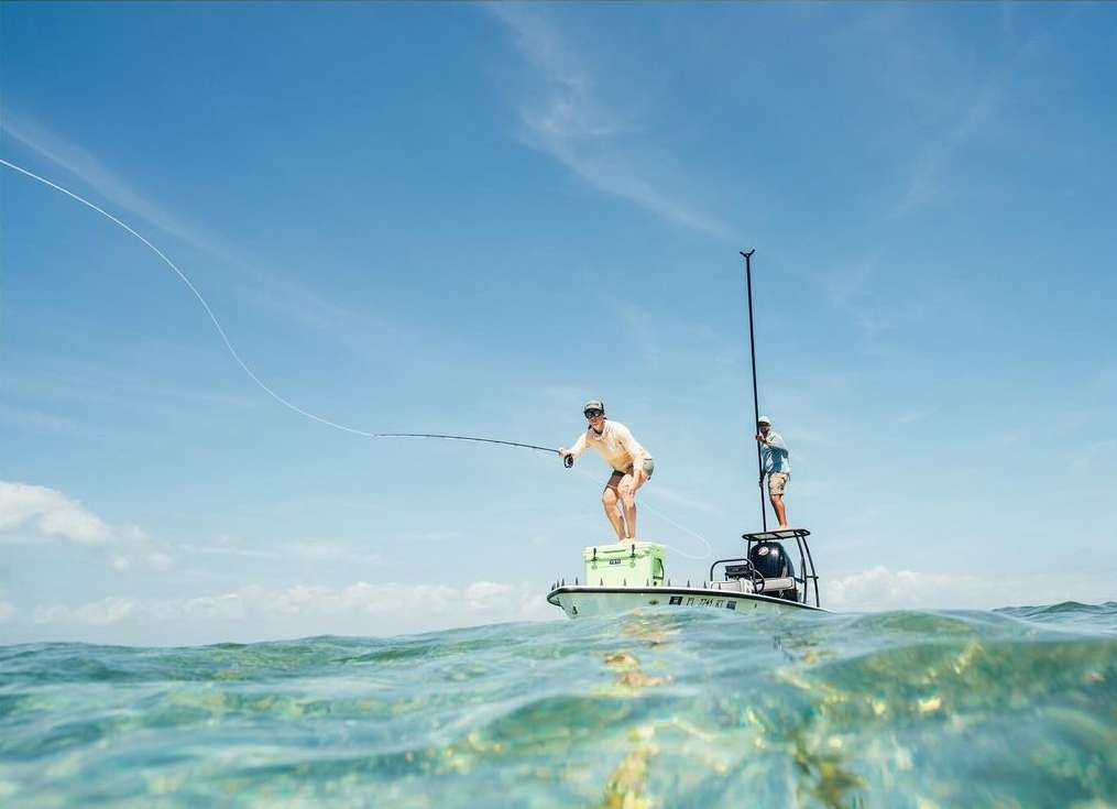 Two adult males are fishing from a boat on clear water with a blue sky backdrop, capturing an outdoor sport scene with active participants.