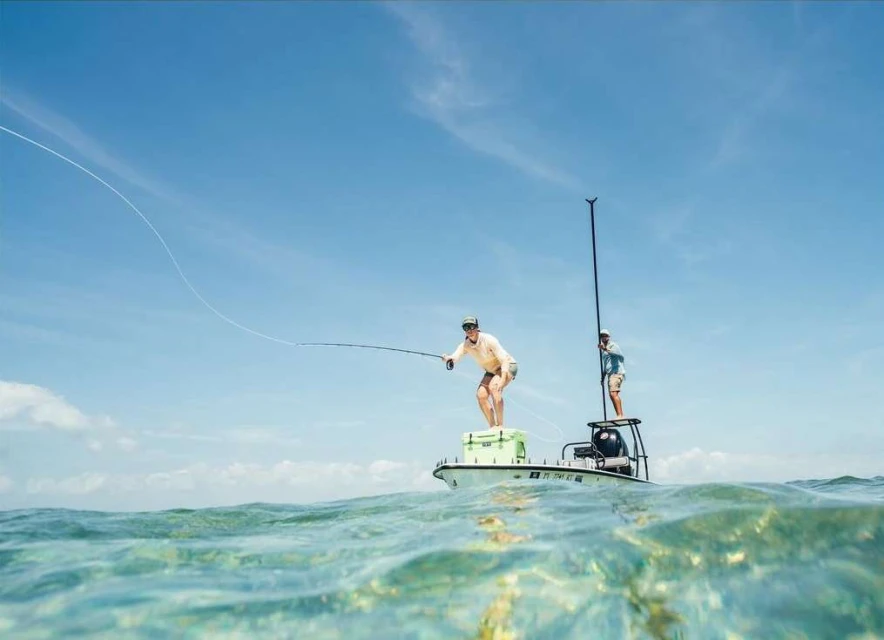Two adult males are fishing from a boat on clear water with a blue sky backdrop, capturing an outdoor sport scene with active participants.