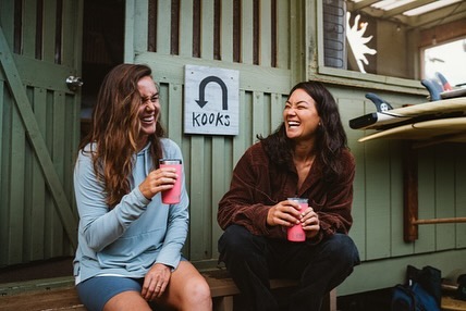 Two women laugh while holding drinks outside a building with surfboards. They are sitting on a bench on a sunny day.