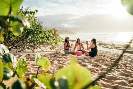 Three women sitting together on a sandy beach at sunset, laughing and holding drinks near the ocean shore.