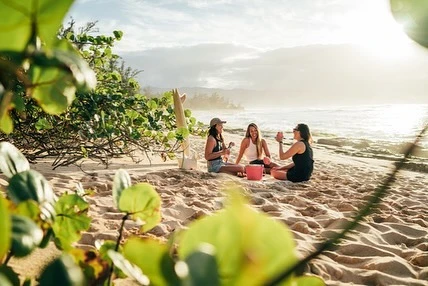 Three women sitting together on a sandy beach at sunset, laughing and holding drinks near the ocean shore.