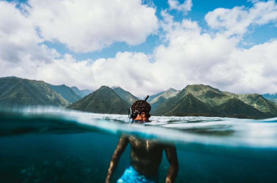 Man swimming underwater with snorkel and mountains in background. Person is partially submerged in ocean with cloudy sky above.