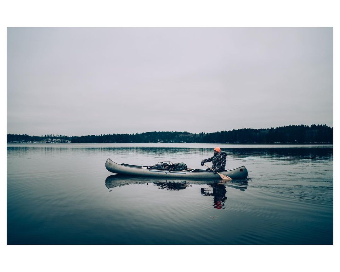 Man in a canoe on a calm lake with forested shoreline under an overcast sky, wearing an orange hat and camouflage jacket.