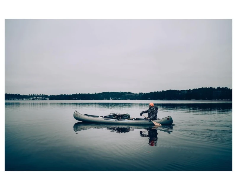 Man in a canoe on a calm lake with forested shoreline under an overcast sky, wearing an orange hat and camouflage jacket.