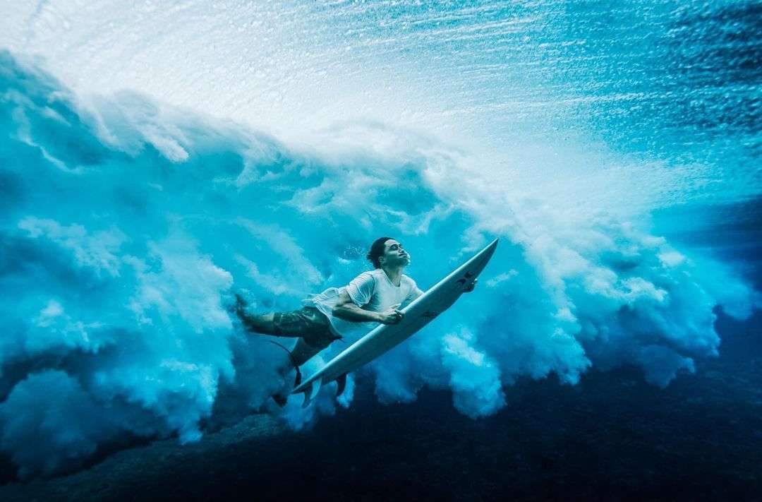 Underwater shot of a surfer in a white t-shirt holding a surfboard beneath a wave, showcasing the power and beauty of ocean surfing.