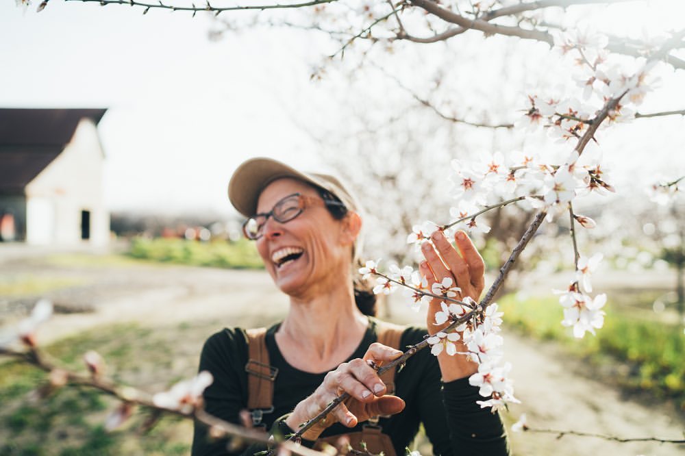Smiling woman in glasses touches almond blossoms on a sunny day with a farm building in the blurred background, capturing springtime beauty.