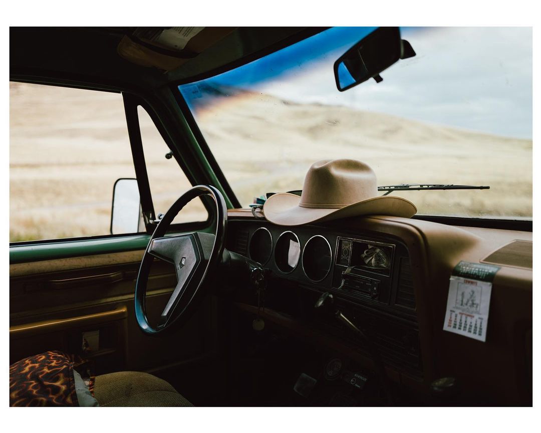 Interior view of an old pickup truck with a cowboy hat on the dashboard, overlooking a tan, grassy field on a cloudy day.