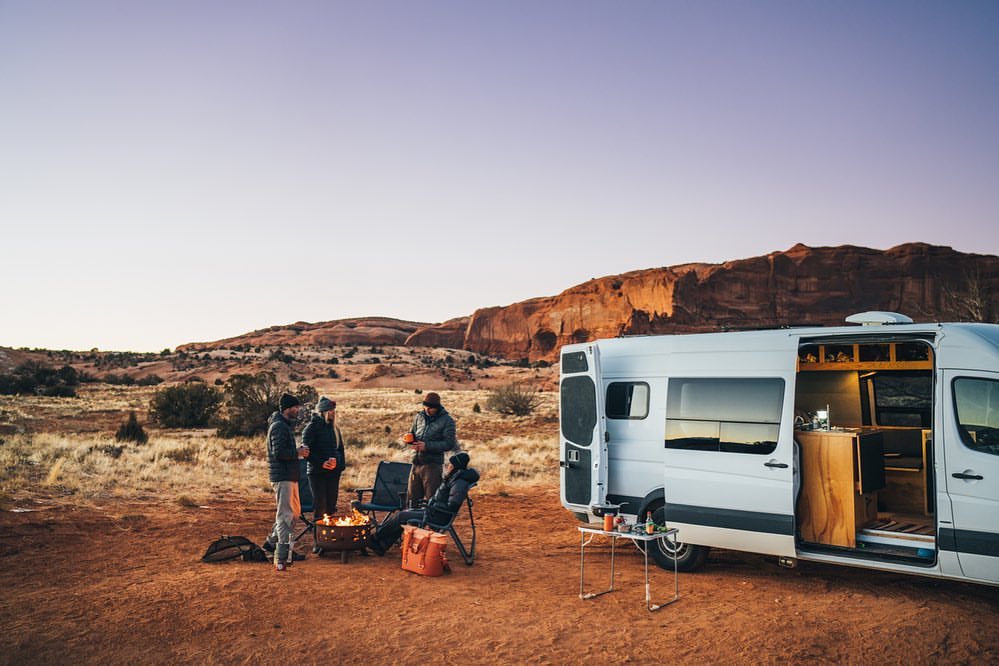 Four people gather around a campfire near a white camper van in a desert landscape at sunset with mountains in the background.