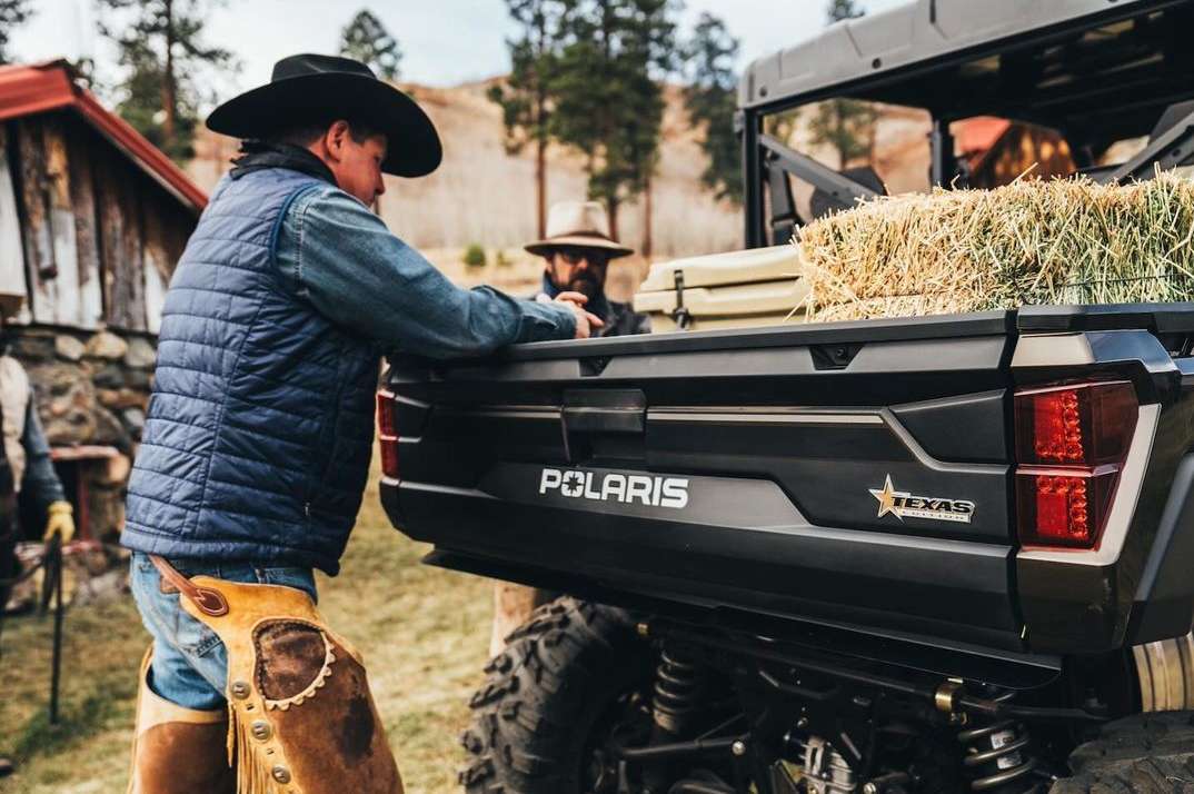 Cowboy near a black Polaris vehicle with hay bales in the back, rural setting, wearing chaps, vest, and hat, with farm buildings in the background.