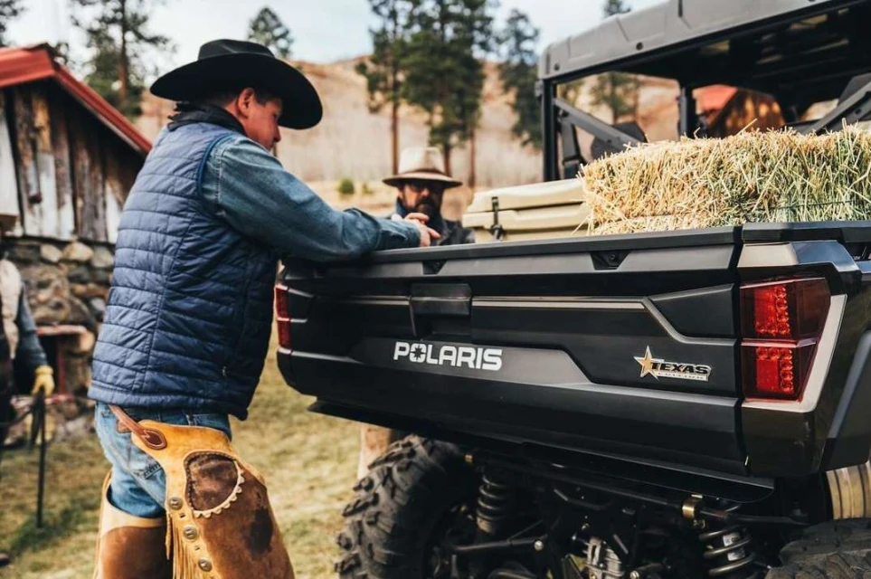 Cowboy near a black Polaris vehicle with hay bales in the back, rural setting, wearing chaps, vest, and hat, with farm buildings in the background.