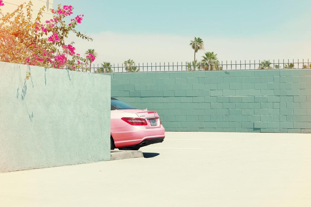 Pink car parked near a light blue brick wall with flowering bougainvillea and palm trees in the background on a bright sunny day.