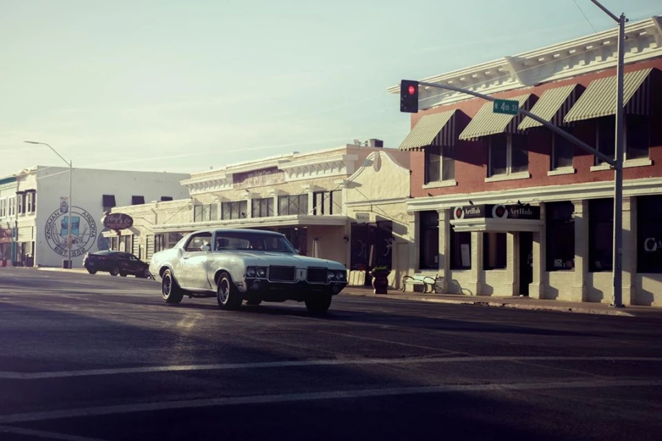 Classic silver Oldsmobile Cutlass driving down main street in small town, Arizona, with buildings and shops in the background on a sunny day.