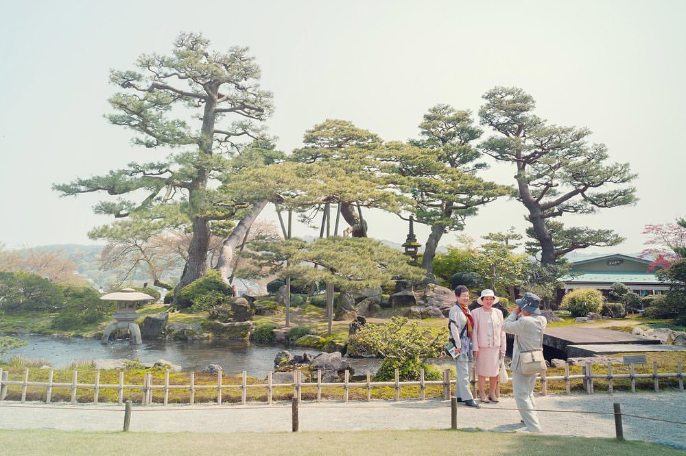 Three people pose in a sunny Japanese garden near a pond and manicured trees, with one adult male photographing two adult females.