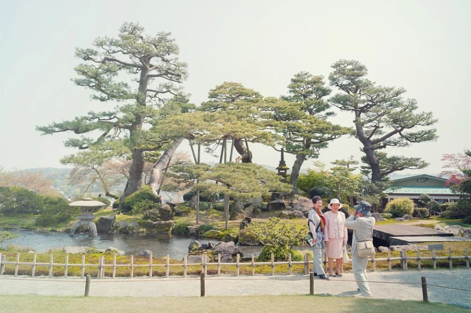 Three people pose in a sunny Japanese garden near a pond and manicured trees, with one adult male photographing two adult females.