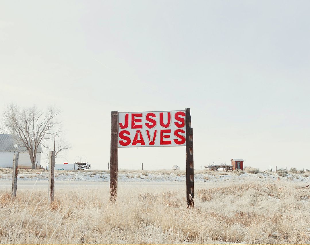 Sign reading "Jesus Saves" in red letters stands in a field of dry grass, with a white building and trailer visible in the distance on a bright day.