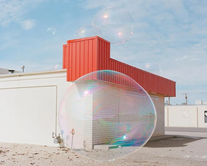 Large iridescent soap bubbles float in front of an abandoned building under a bright blue sky, creating a surreal outdoor scene.