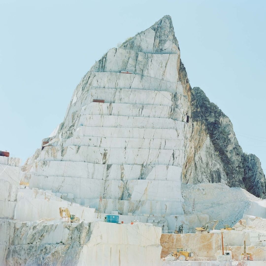Marble quarry featuring stepped terraces of white stone forming a large mountain-like peak against a clear sky. Quarry equipment visible.