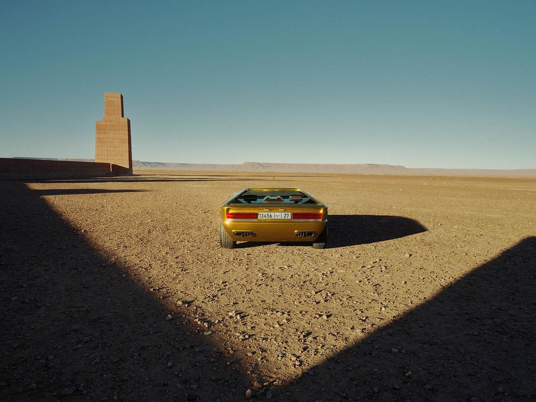 Rear view of a gold sportscar parked in a desert landscape under a clear blue sky, casting shadows on the ground.