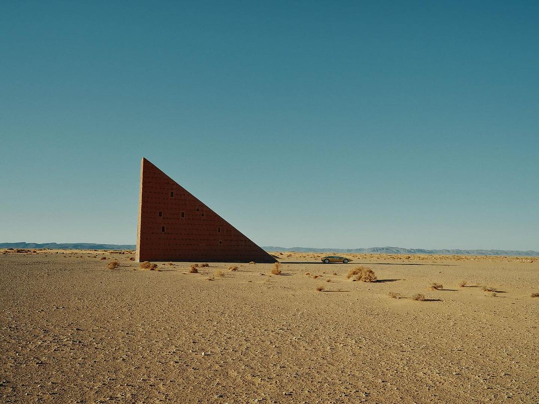 Architectural art structure in desert landscape, clear blue sky background, featuring desert vegetation and distant mountains.