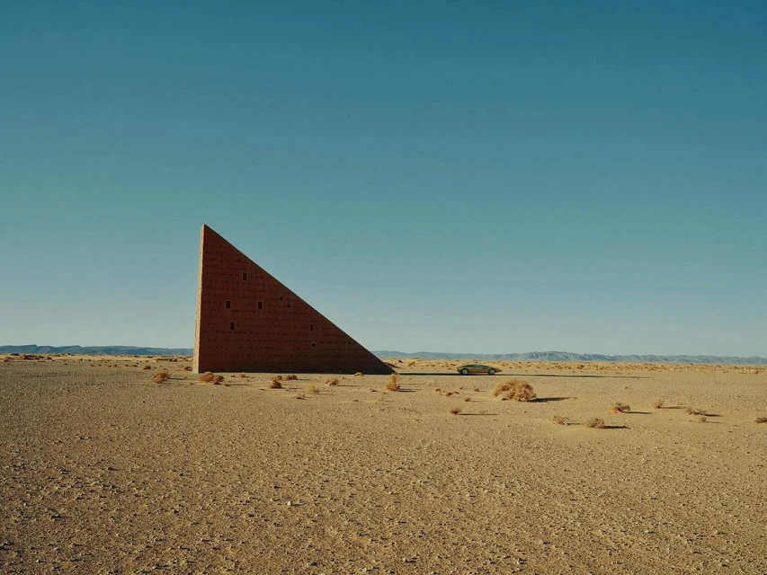 Architectural art structure in desert landscape, clear blue sky background, featuring desert vegetation and distant mountains.