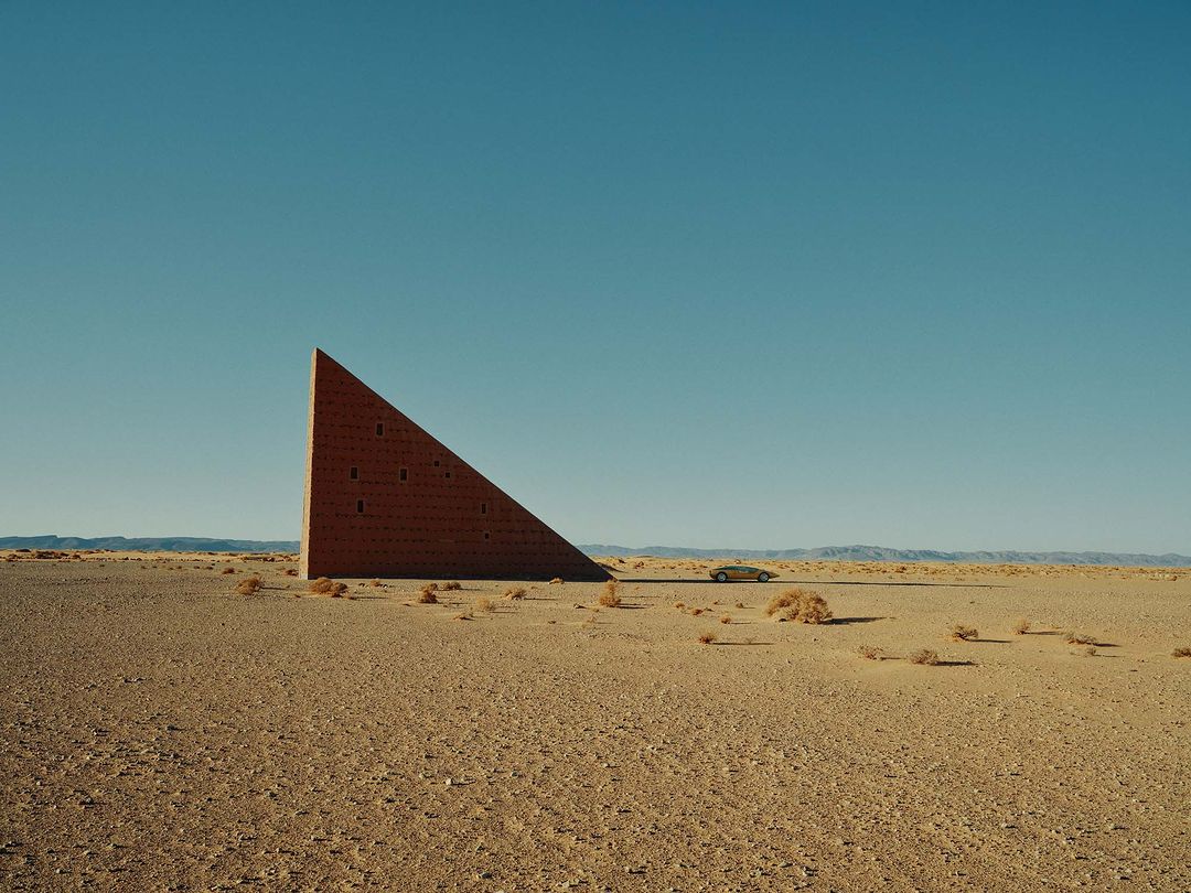 Minimalist shot of a brick triangular building in a sandy desert with a distant car under a clear blue sky, creating a surreal landscape.