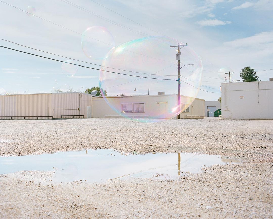 Large iridescent soap bubbles float in a desolate gravel lot with white buildings under a blue sky, reflecting in a puddle.