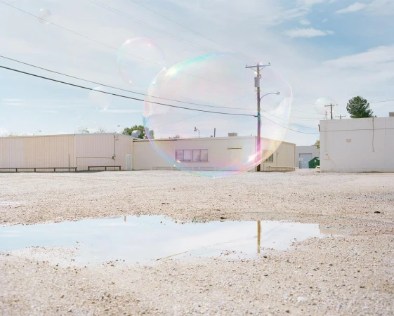 Large iridescent soap bubbles float in a desolate gravel lot with white buildings under a blue sky, reflecting in a puddle.