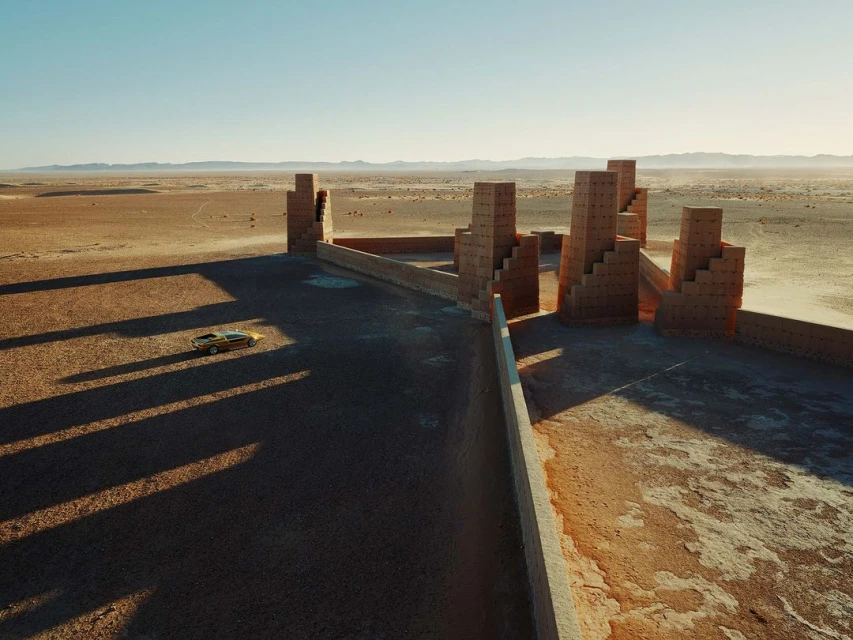 Aerial shot of a classic yellow car in a desert landscape near unique brick architecture, under a clear sky, creating a surreal scene.