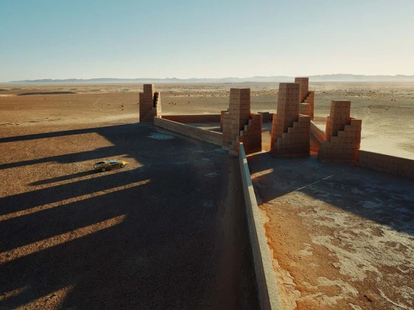 Aerial shot of a classic yellow car in a desert landscape near unique brick architecture, under a clear sky, creating a surreal scene.
