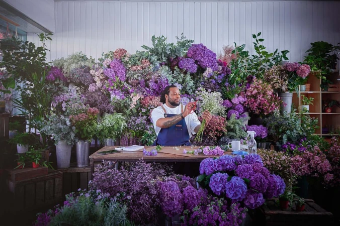 Man arranges flowers in a lush indoor flower shop filled with purple hydrangeas, greenery, and blooms, on a wooden table.