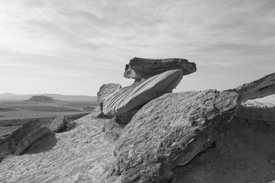 Black and white shot of a rock formation with a cloth draped over it, set against a desolate landscape and cloudy sky.