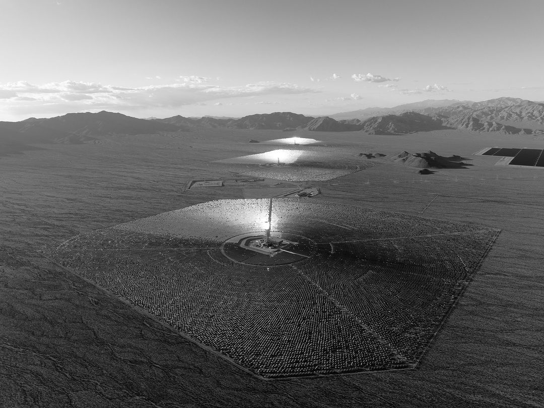 Black and white aerial view of a solar power plant in a desert landscape with rows of heliostats and a central receiver tower.