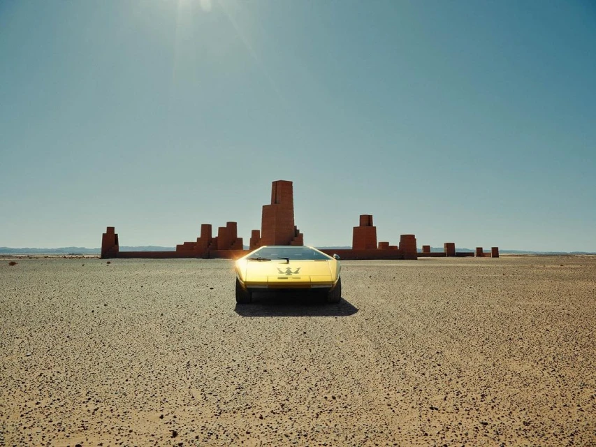 Yellow vintage Maserati Merak car in desert landscape with geometric brick architecture under clear blue sky and bright sunlight.