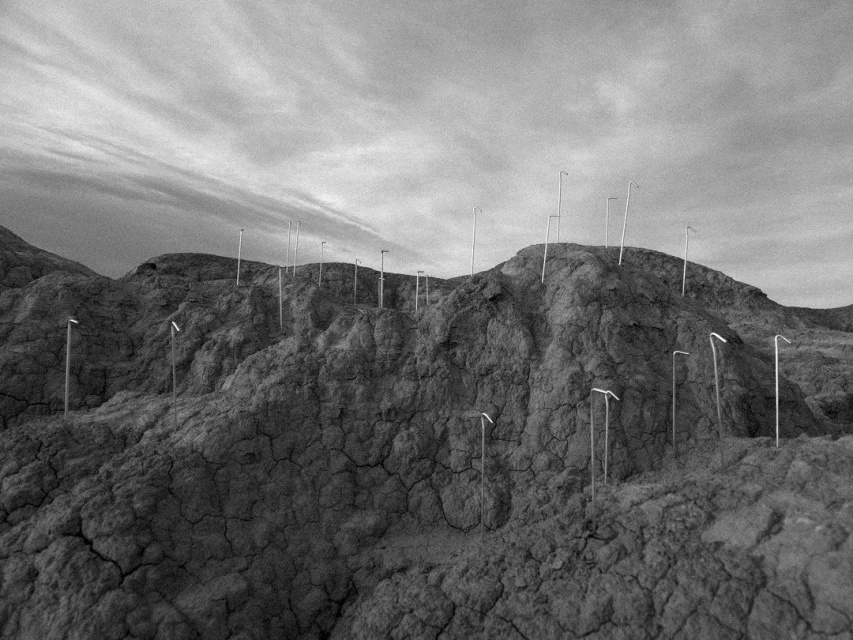 Monochrome landscape featuring a cracked mud hill covered in drinking straws, under a cloudy sky, creating an eerie, abstract outdoor scene.