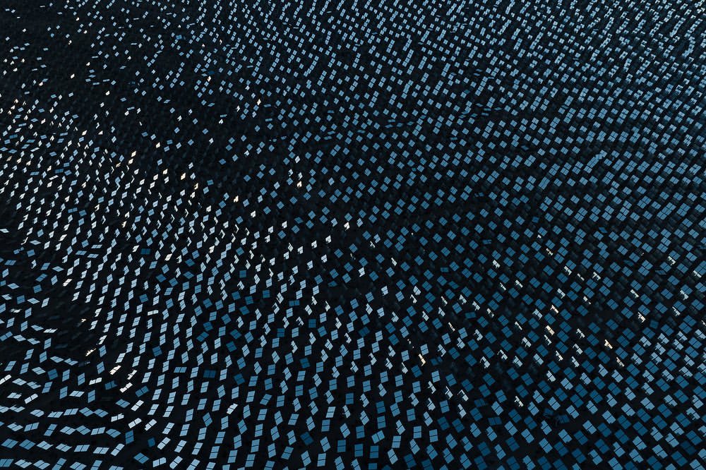 High-angle view of a solar panel field with many square panels, creating a patterned landscape of blues and dark hues.