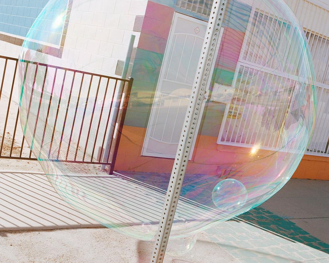 Large transparent soap bubble floating outdoors, reflecting the surrounding building, fence, and sky with pastel iridescence.