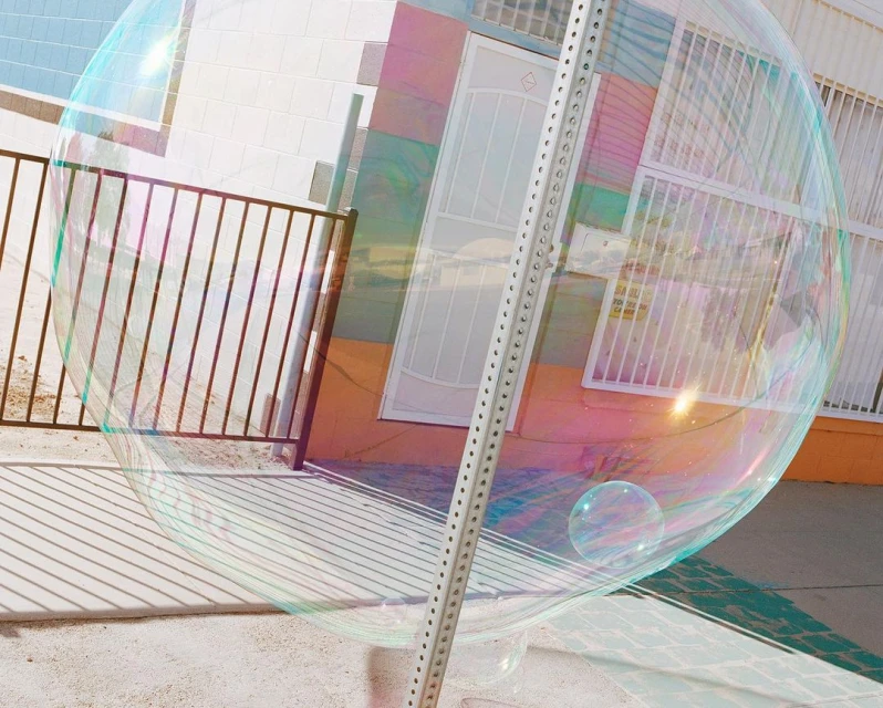 Large transparent soap bubble floating outdoors, reflecting the surrounding building, fence, and sky with pastel iridescence.