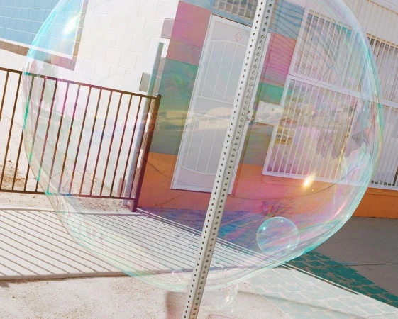 Large transparent soap bubble floating outdoors, reflecting the surrounding building, fence, and sky with pastel iridescence.