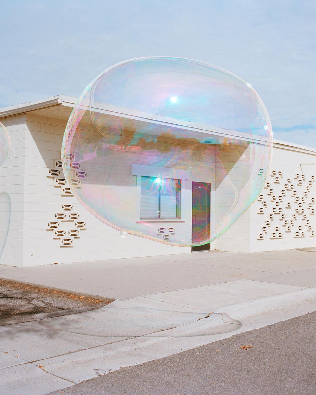 A large soap bubble hovers in front of a one-story white building with a decorative block pattern on a sunny day in an outdoor environment.