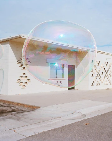 A large soap bubble hovers in front of a one-story white building with a decorative block pattern on a sunny day in an outdoor environment.