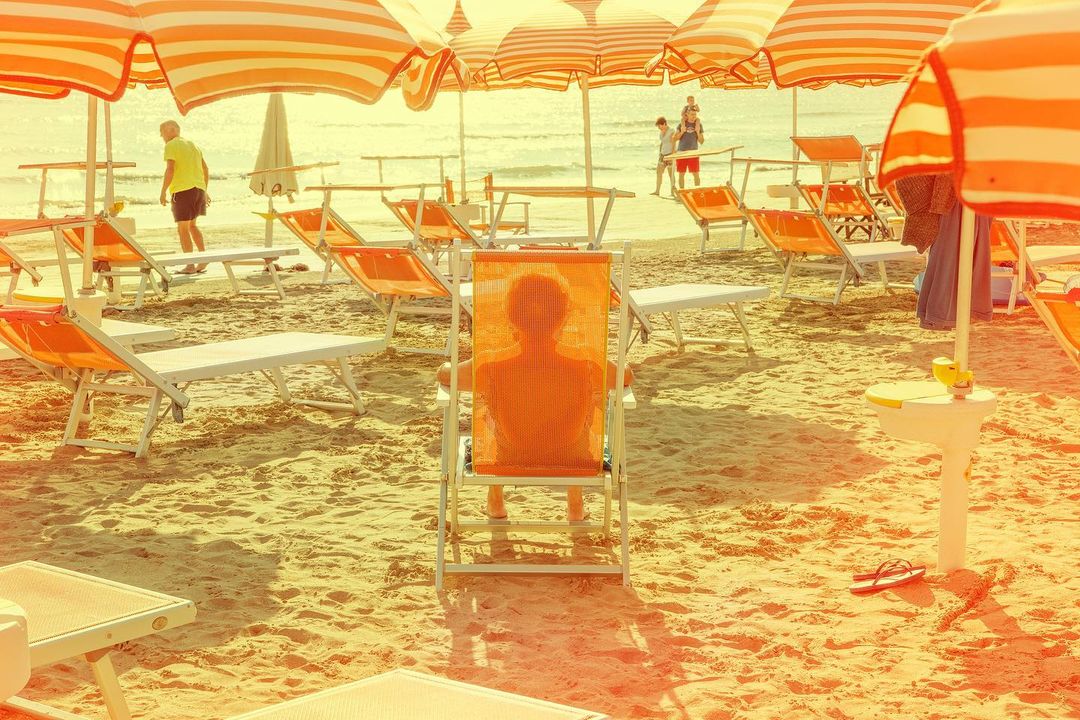 Beach scene with person sitting in orange deck chair under striped umbrella.  Sandy beach and calm ocean in background.