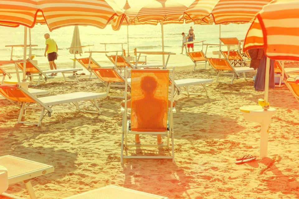 Beach scene with person sitting in orange deck chair under striped umbrella.  Sandy beach and calm ocean in background.